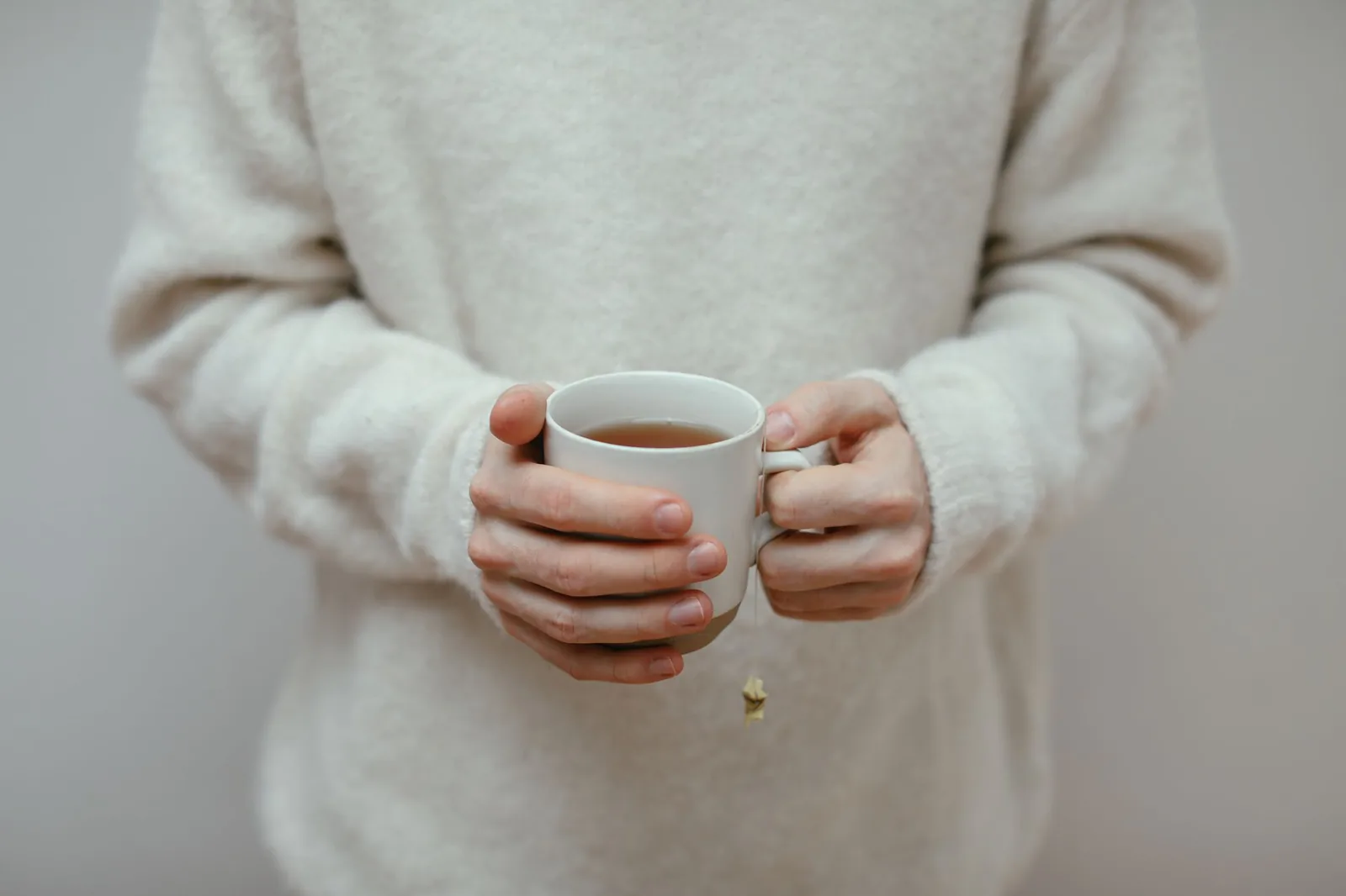 Hands holding a small cup of tea in front of a cream jumper