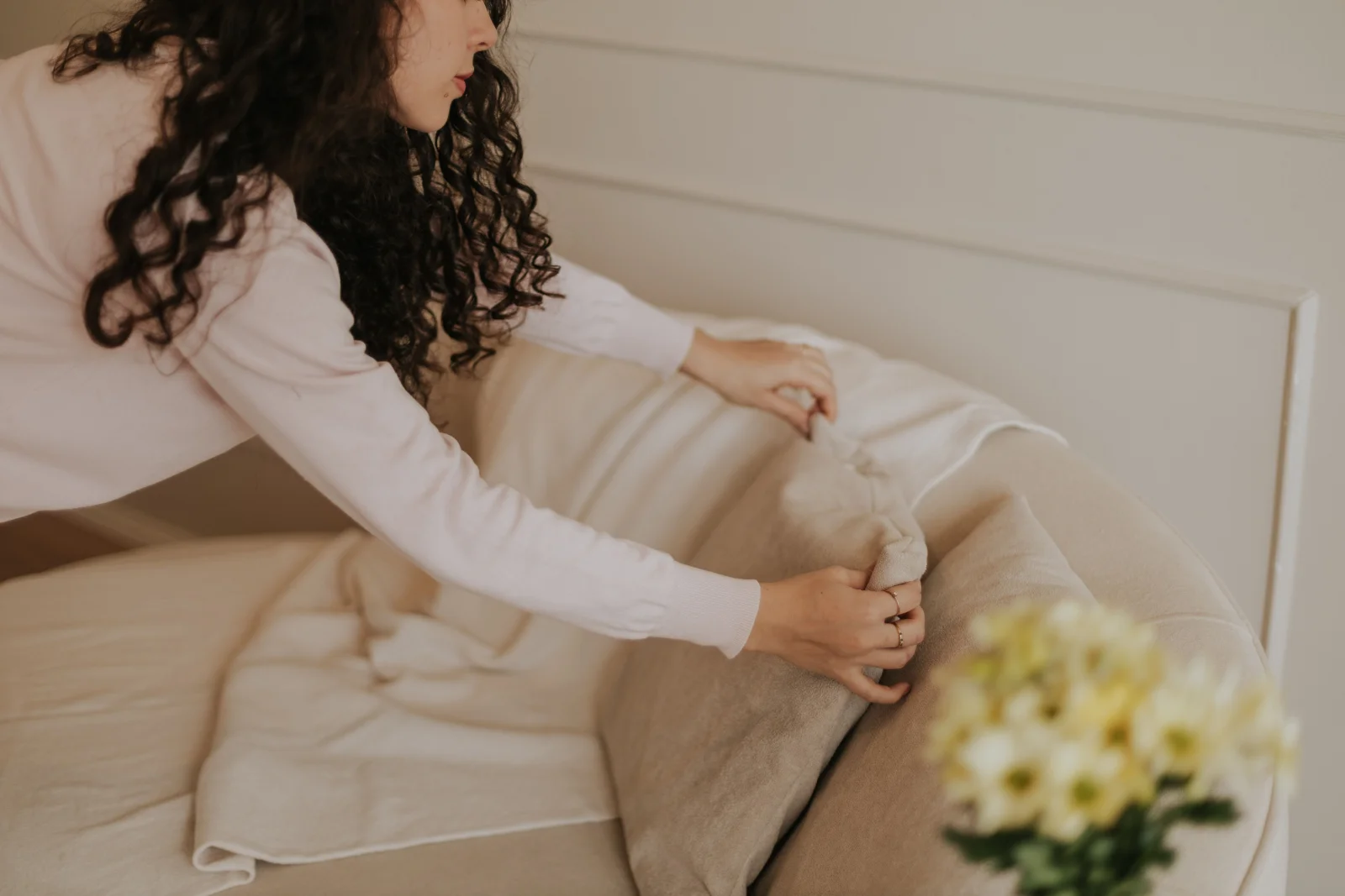 A woman in a long-sleeved top arranging cushions on a cream sofa in a bright living room